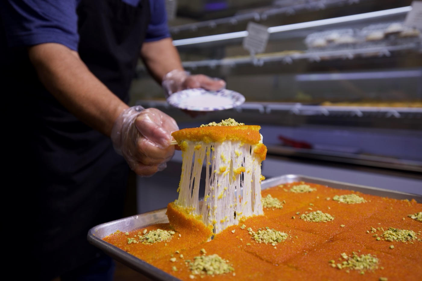 Freshly made Knafeh Nabulsieh with golden syrup being poured and a dramatic cheese pull, topped with pistachios